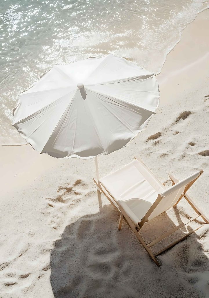 A single white beach chair and parasol on a sunlit sandy beach beside calm turquoise water, symbolising rest, space and clarity for business owners seeking balance